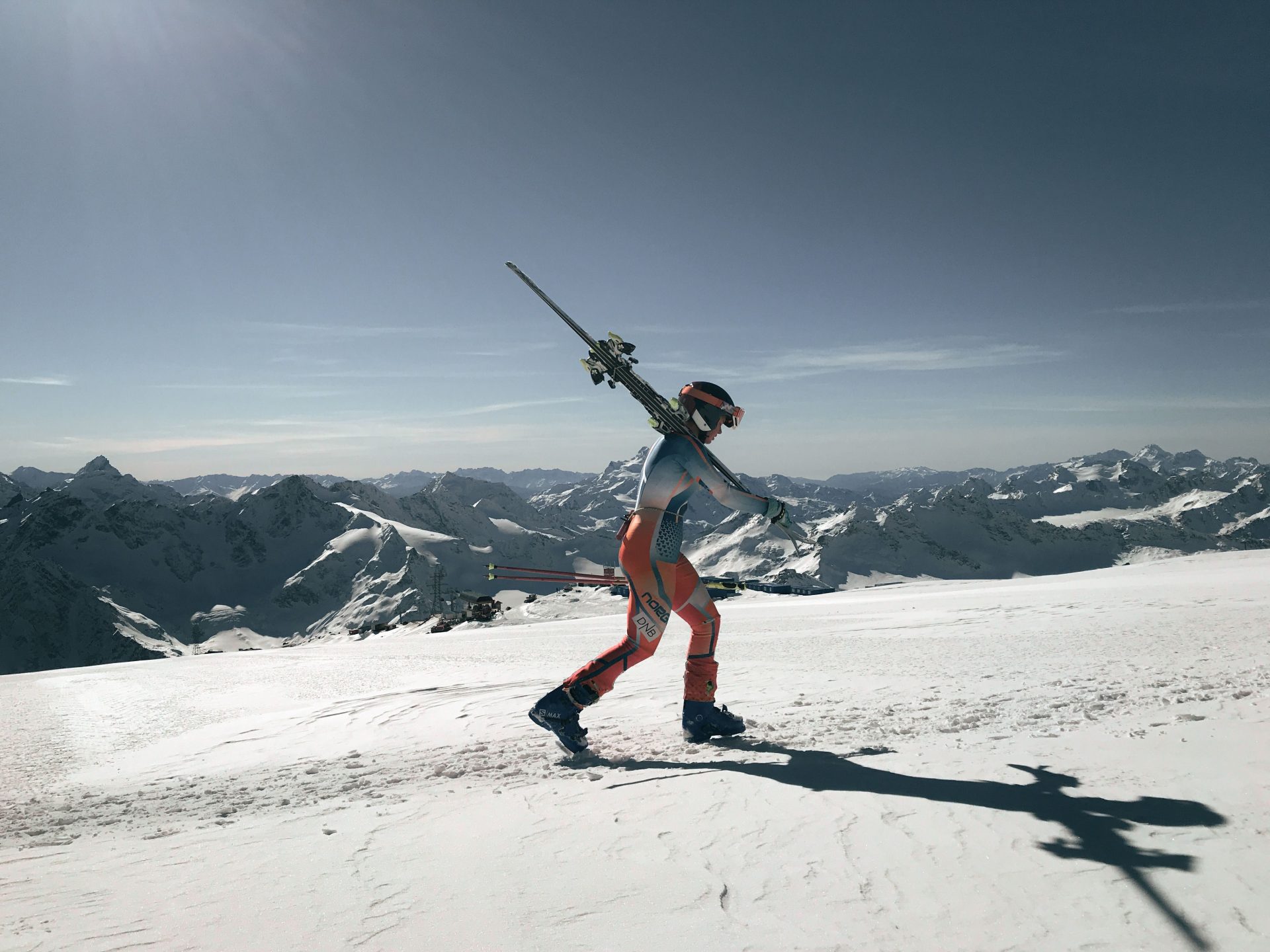 A skier carrying skis on a snowy mountain with a panoramic view of the Alps under a clear sky.