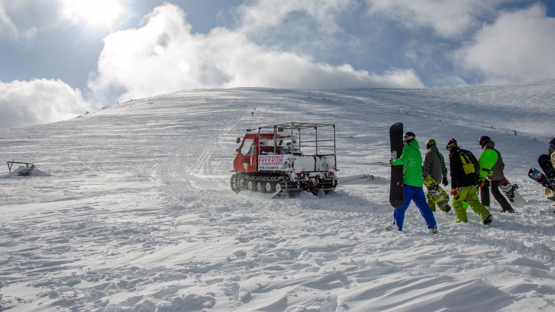 Snowboarders walking near a snow groomer on a snowy hill in winter sunshine.