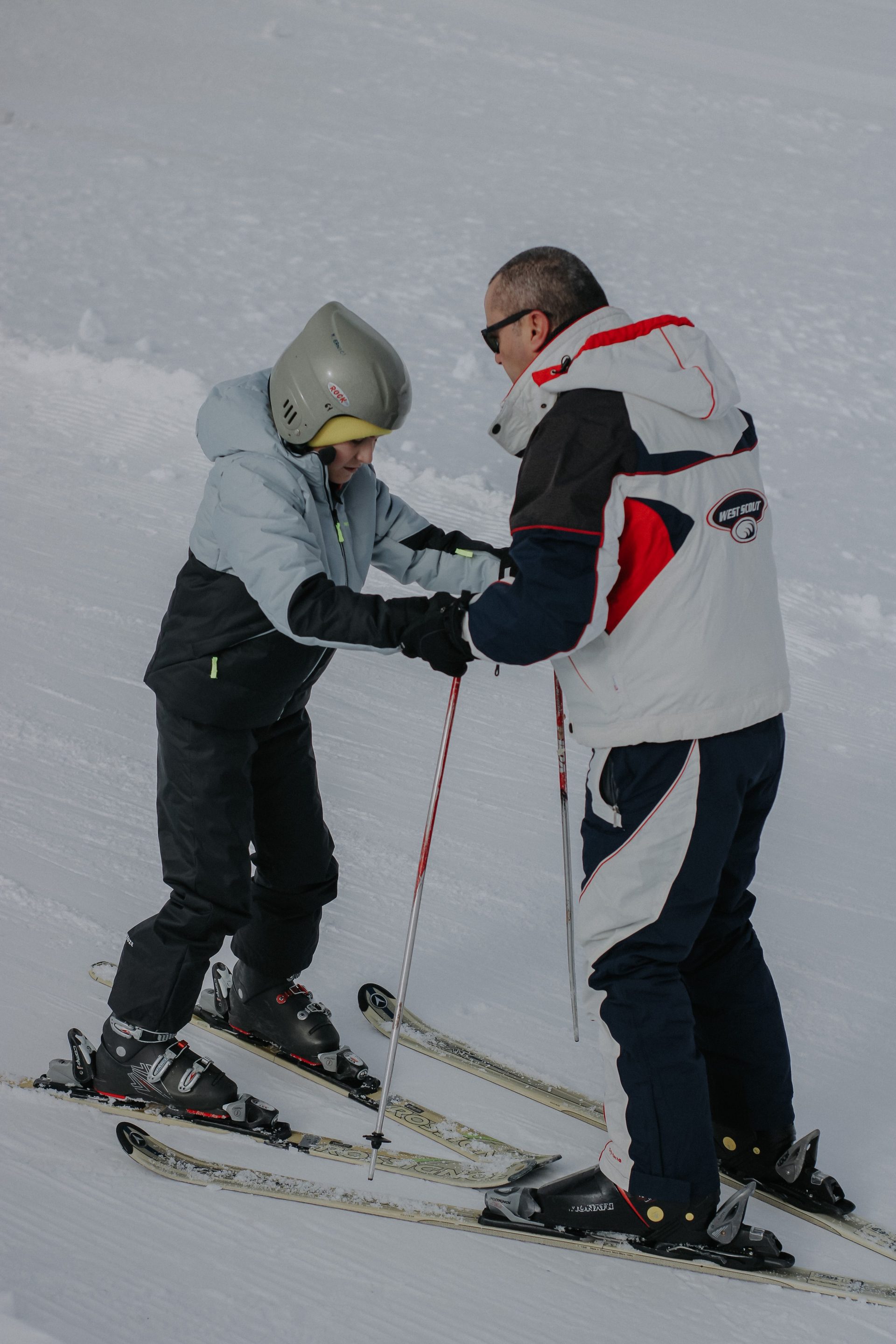 A father helps his child learn to ski on a snowy slope, wearing warm winter gear.