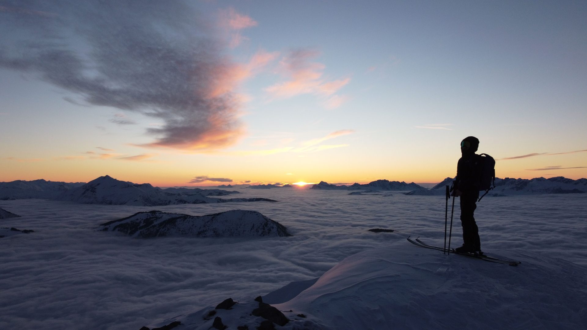 A lone skier stands atop a snowy peak in Les Houches, overlooking a stunning sunset.